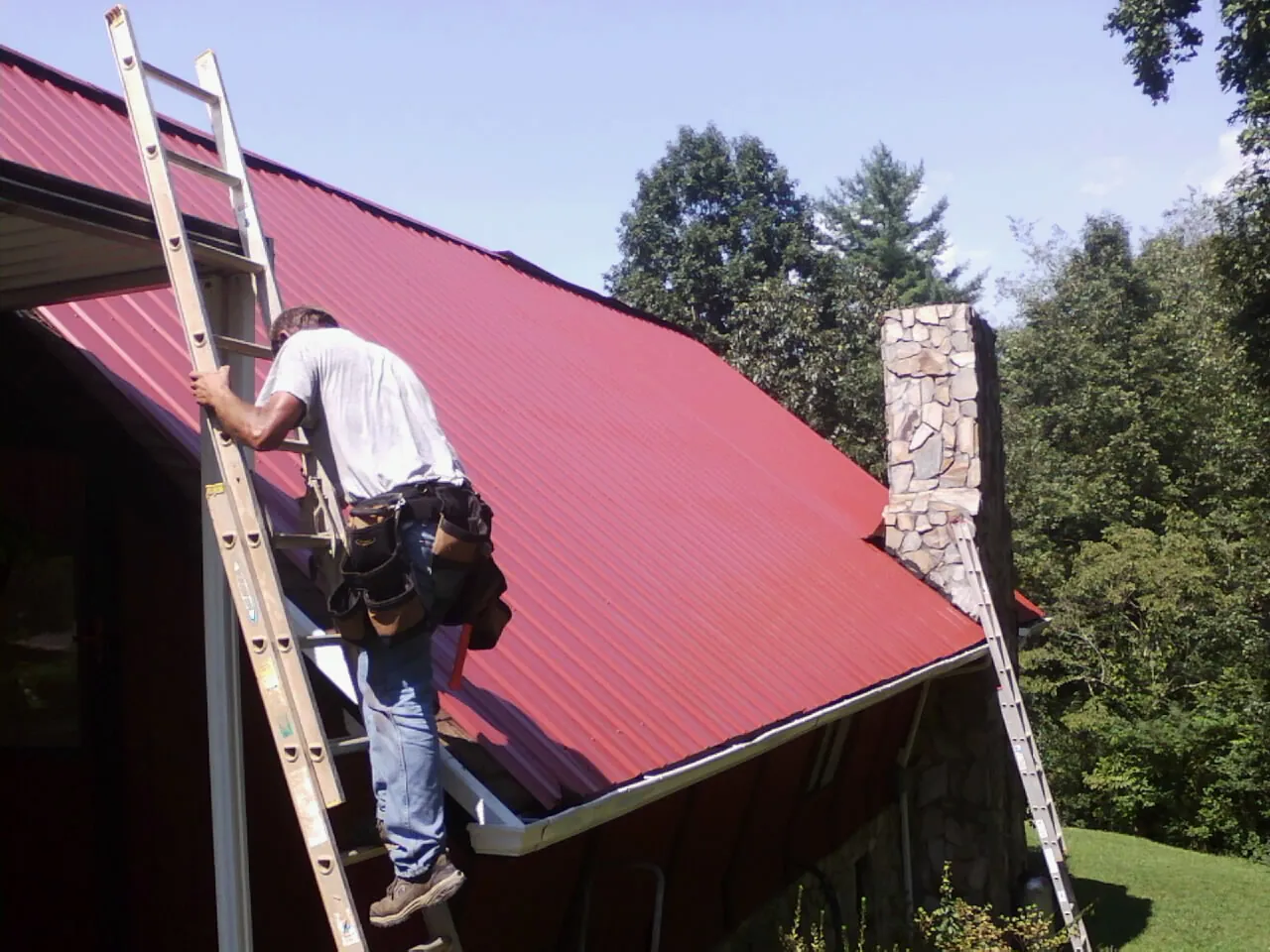 Roofer on a ladder beside a red metal roof and stone chimney — Soffit & Fascia Repair by Roofing Brothers in South Fork